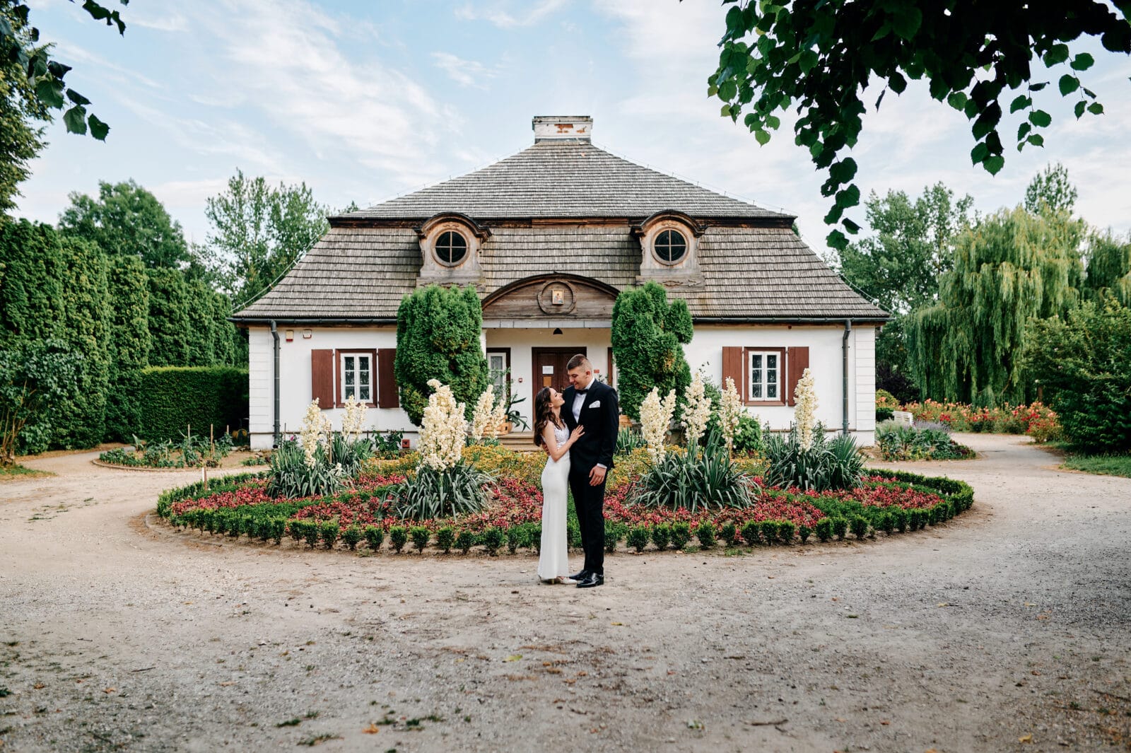 Naturalne ujęcie pary na tle drewnianych budynków Muzeum Wsi Lubelskiej, podkreślające ich elegancję i wyjątkowy klimat tego miejsca.